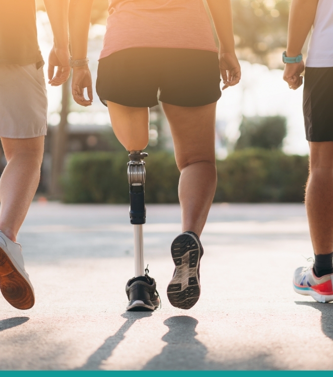 Person with a prosthetic leg walks outdoors alongside two companions in warm sunlight.