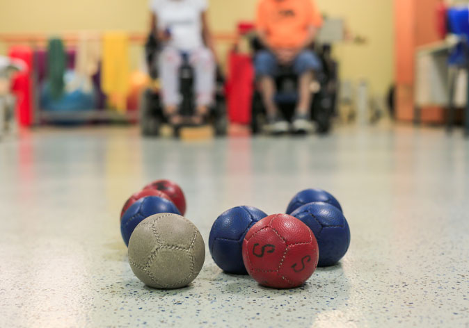Red, blue, and white boccia balls clustered on an indoor court, with two wheelchair players in the background.