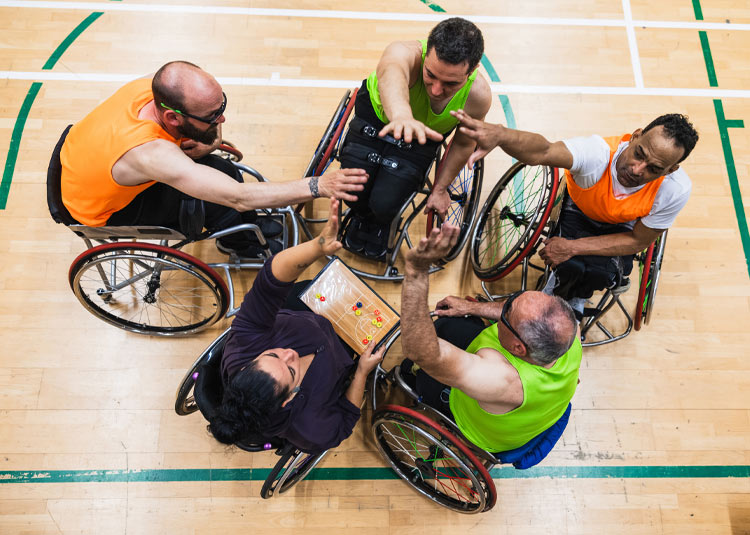 Five wheelchair athletes gather in a circle on an indoor court, reaching toward the centre in a team huddle.