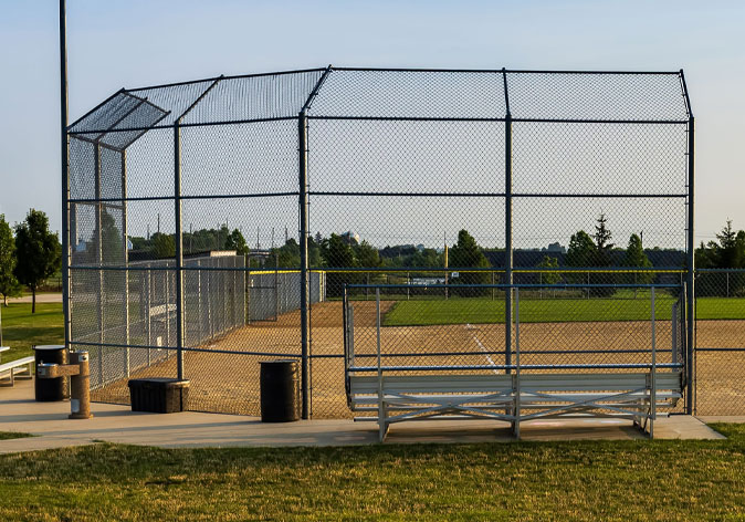 Outdoor baseball diamond enclosed by a chain-link backstop, with empty bleachers beside the field.