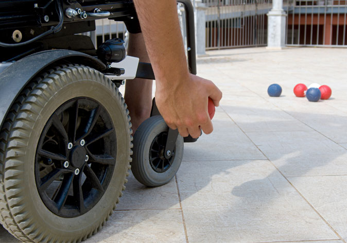 A close-up view focuses on a person in a motorized wheelchair preparing to throw a red boccia ball on an outdoor tiled court.