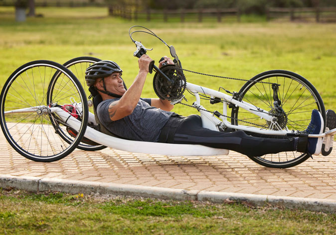 A man wearing a helmet rides a recumbent handcycle bike along a paved brick path in an outdoor park.