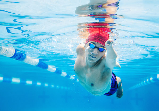 An underwater shot captures a para-swimmer wearing a red cap and goggles gliding through a bright blue pool lane.