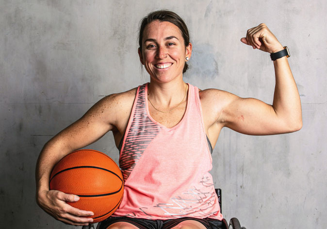 A smiling female wheelchair basketball player flexes her left bicep while holding a basketball under her right arm against a textured gray wall.