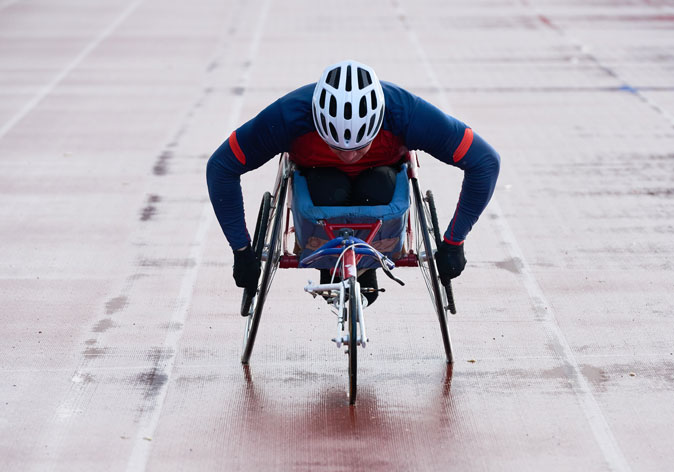 A wheelchair racer wearing a white helmet and blue long-sleeve shirt leans forward while competing on a running track.