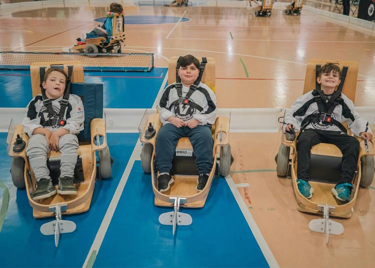 Three children seated in adaptive power chairs on an indoor sports court, participating in an inclusive recreation program.
